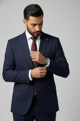 Man adjusting navy two-piece slim fit suit with peak lapel and burgundy tie.