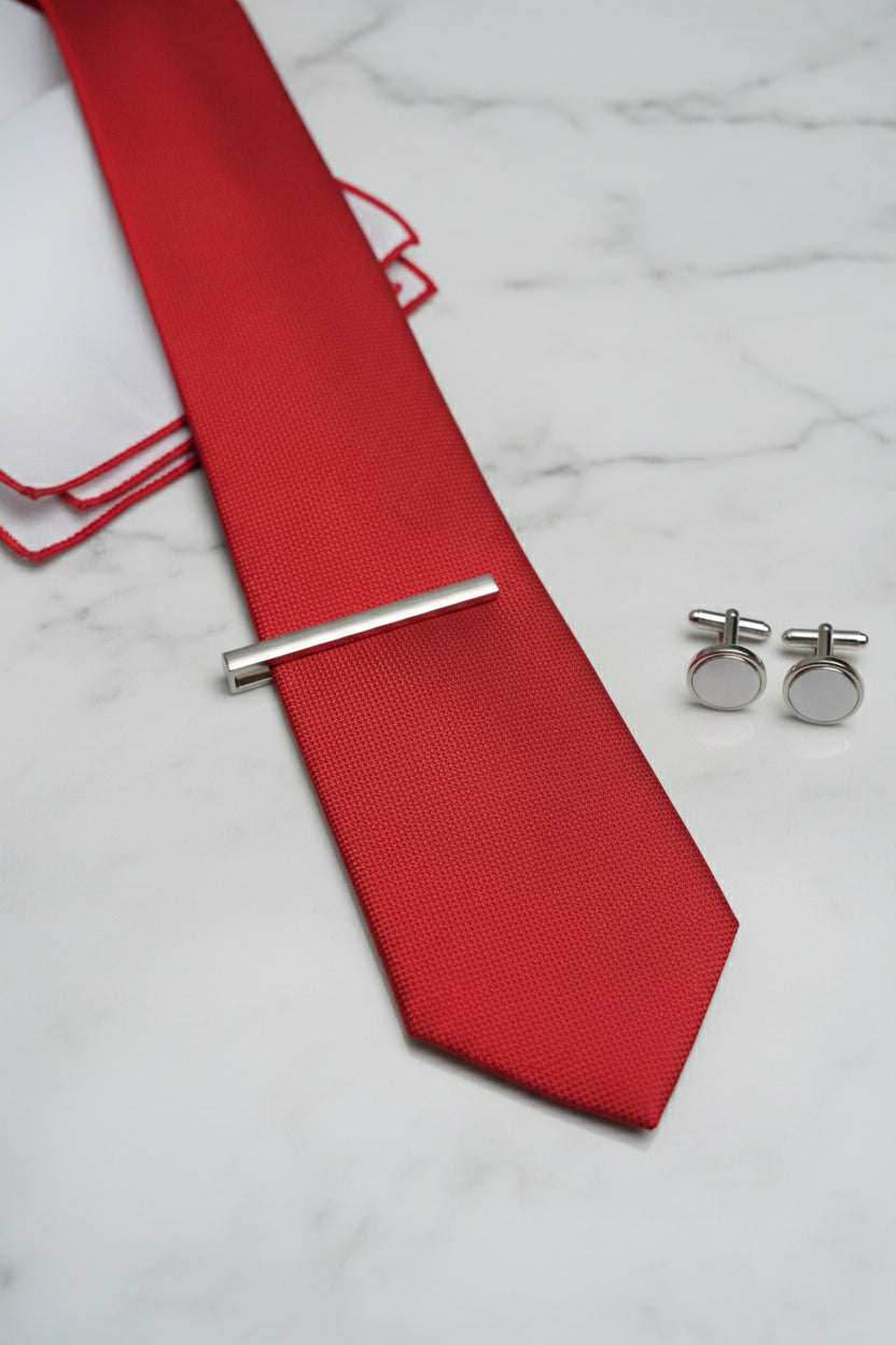 Red textured silk tie displayed with silver tie clip and cufflinks on marble surface.